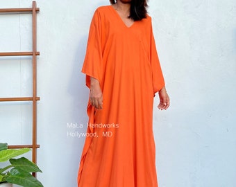 Puede incluir: Un vestido caftán naranja, largo hasta el suelo, con cuello en V y mangas anchas. El vestido está hecho de una tela ligera y tiene un corte holgado. El texto "MaLa Handworks Hollywood, MD" es visible en el vestido.
