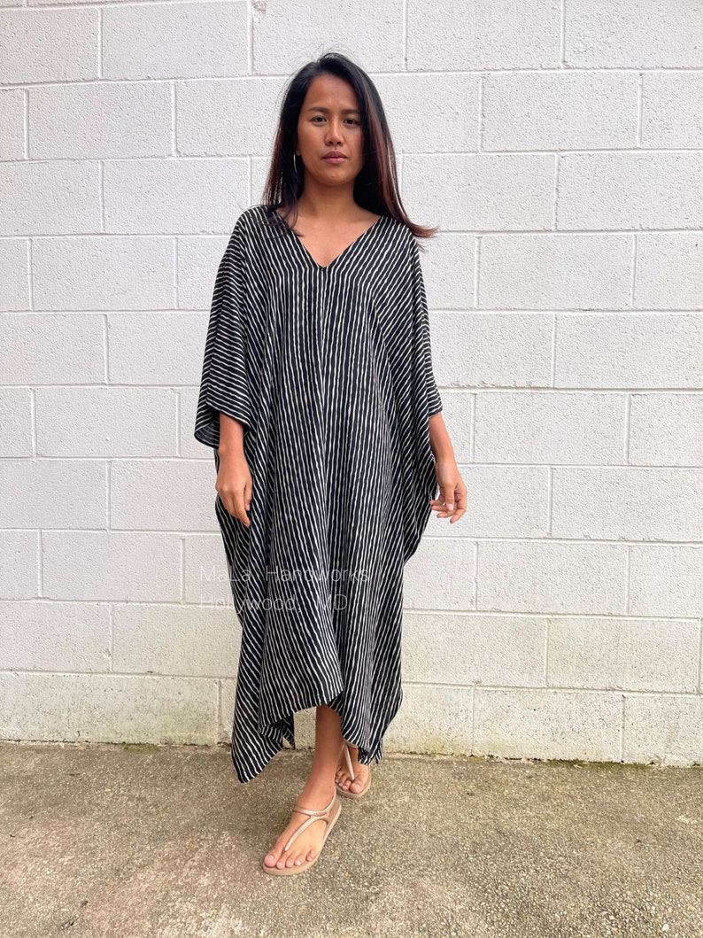 K&ouml;nnte beinhalten: Ein schwarz-wei&szlig; gestreiftes Kaftan mit V-Ausschnitt. Der Kaftan ist aus einem leichten Stoff gefertigt und hat eine flie&szlig;ende Silhouette. Das Model tr&auml;gt den Kaftan mit Sandalen.