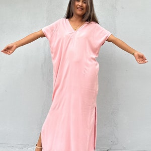 Pode incluir: Um vestido maxi rosa claro, com decote em V, mangas curtas e fendas laterais. O vestido é feito de um tecido macio e fluido. A modelo está usando sandálias de salto alto na cor nude.