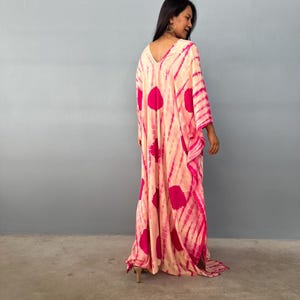 May include: A long, flowing kaftan dress with a V-neckline. The dress has a tie-dye pattern in peach and pink. The design includes large pink spots and stripes. The dress has long sleeves and a relaxed fit.