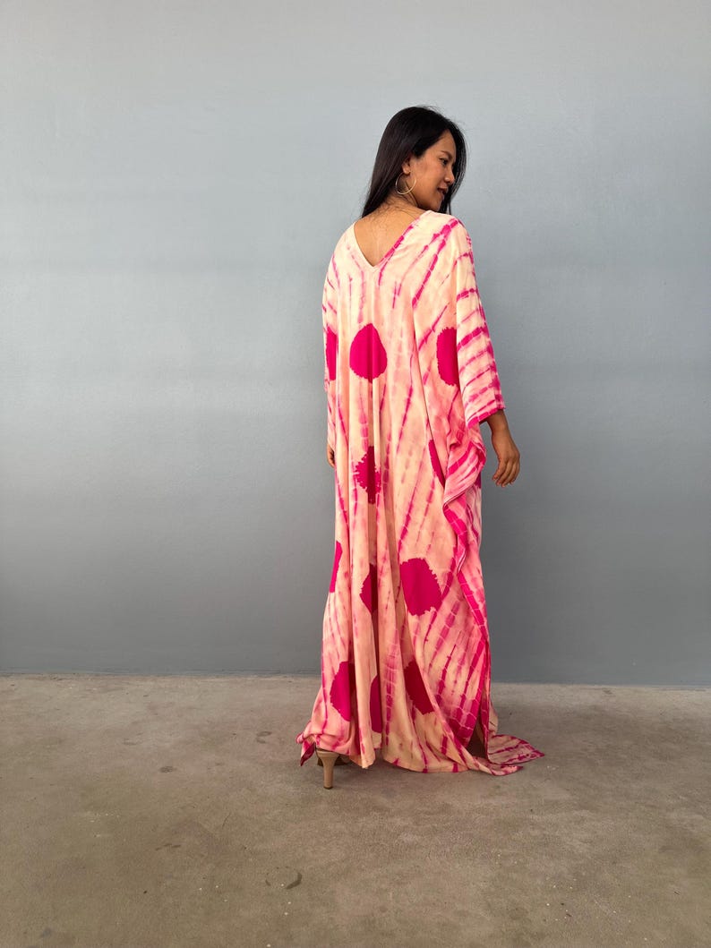 May include: A long, flowing kaftan dress with a V-neckline. The dress has a tie-dye pattern in peach and pink. The design includes large pink spots and stripes. The dress has long sleeves and a relaxed fit.