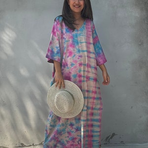 Op de afbeelding: Een lange, vloeiende tie-dye jurk in tinten roze, blauw en wit. De jurk heeft een V-hals en een ontspannen pasvorm. Het model houdt een witte strohoed vast.