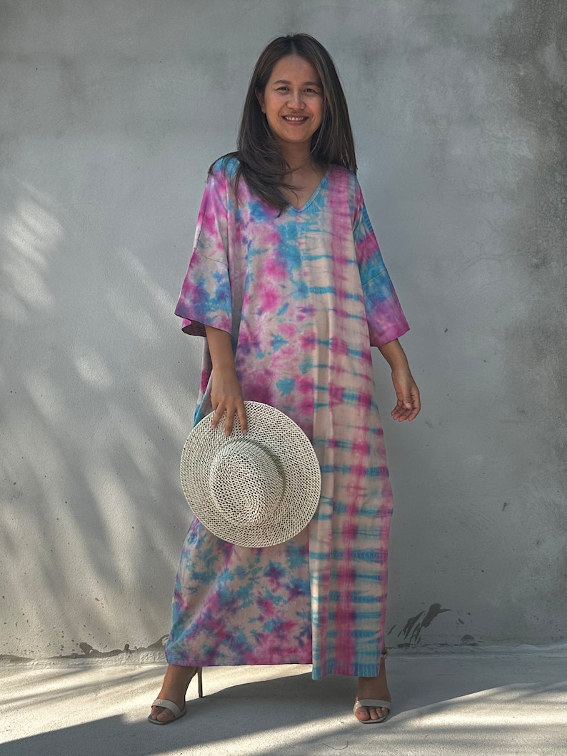 Op de afbeelding: Een lange, vloeiende tie-dye jurk in tinten roze, blauw en wit. De jurk heeft een V-hals en een ontspannen pasvorm. Het model houdt een witte strohoed vast.