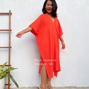 May include: A woman wearing a bright orange, loose-fitting, knee-length dress with a V-neck and side slits. The dress has a textured fabric and the woman is smiling. The text "MALA handworks Hollywood, MD" is visible on the dress.