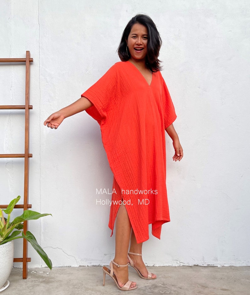 May include: A woman wearing a bright orange, loose-fitting, knee-length dress with a V-neck and side slits. The dress has a textured fabric and the woman is smiling. The text "MALA handworks Hollywood, MD" is visible on the dress.