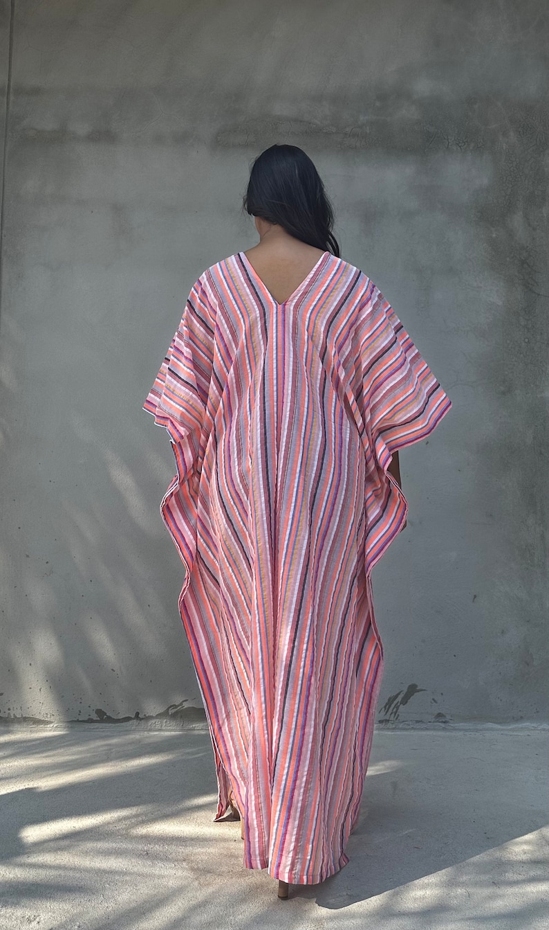 May include: A long, flowing, striped kaftan with a V-neckline. The kaftan is made of a lightweight fabric and has a relaxed fit. The colors of the stripes are pink, orange, white, and gray.