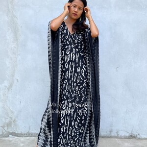 May include: A black and white patterned maxi dress with a V-neckline and long sleeves. The dress has a flowing silhouette and is made of a lightweight fabric. The dress is worn by a person standing in front of a gray wall. The text "Malla Handworks Hollywood MD" is visible on the dress.