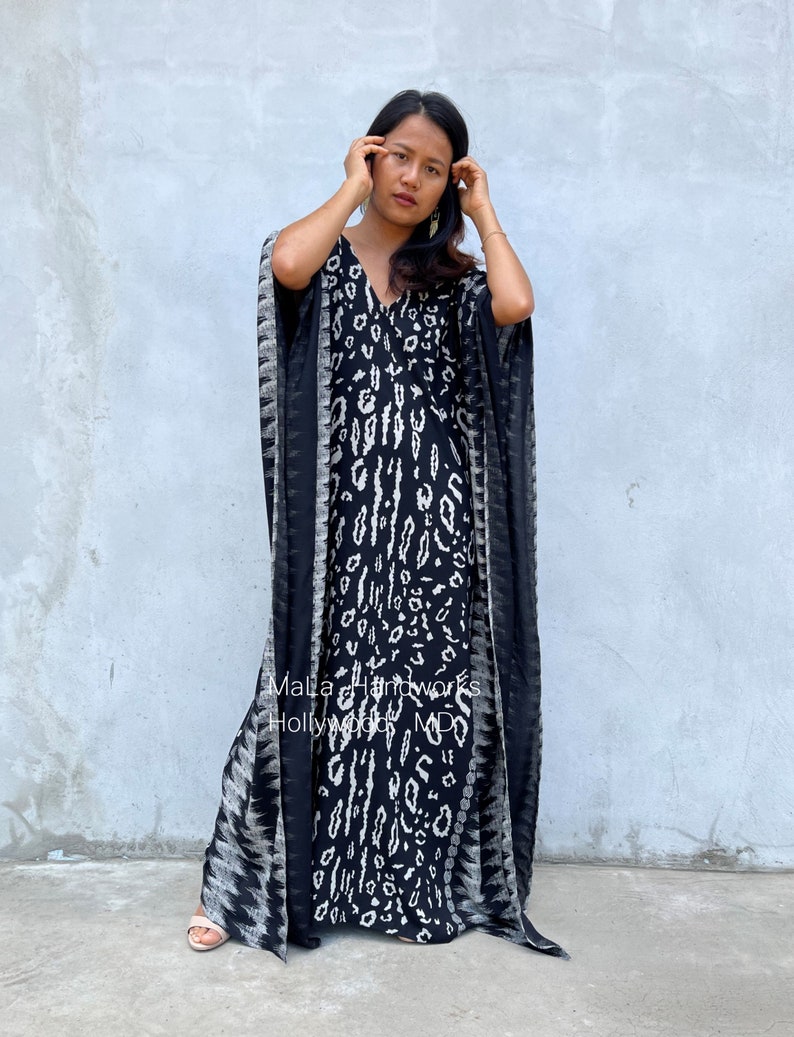 May include: A black and white patterned maxi dress with a V-neckline and long sleeves. The dress has a flowing silhouette and is made of a lightweight fabric. The dress is worn by a person standing in front of a gray wall. The text "Malla Handworks Hollywood MD" is visible on the dress.
