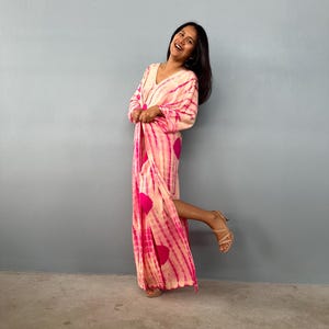May include: A long, flowing dress with a V-neckline, featuring a tie-dye pattern in shades of pink and white. The dress has a vibrant design with large pink circles and vertical stripes. The model is wearing beige high-heeled sandals.