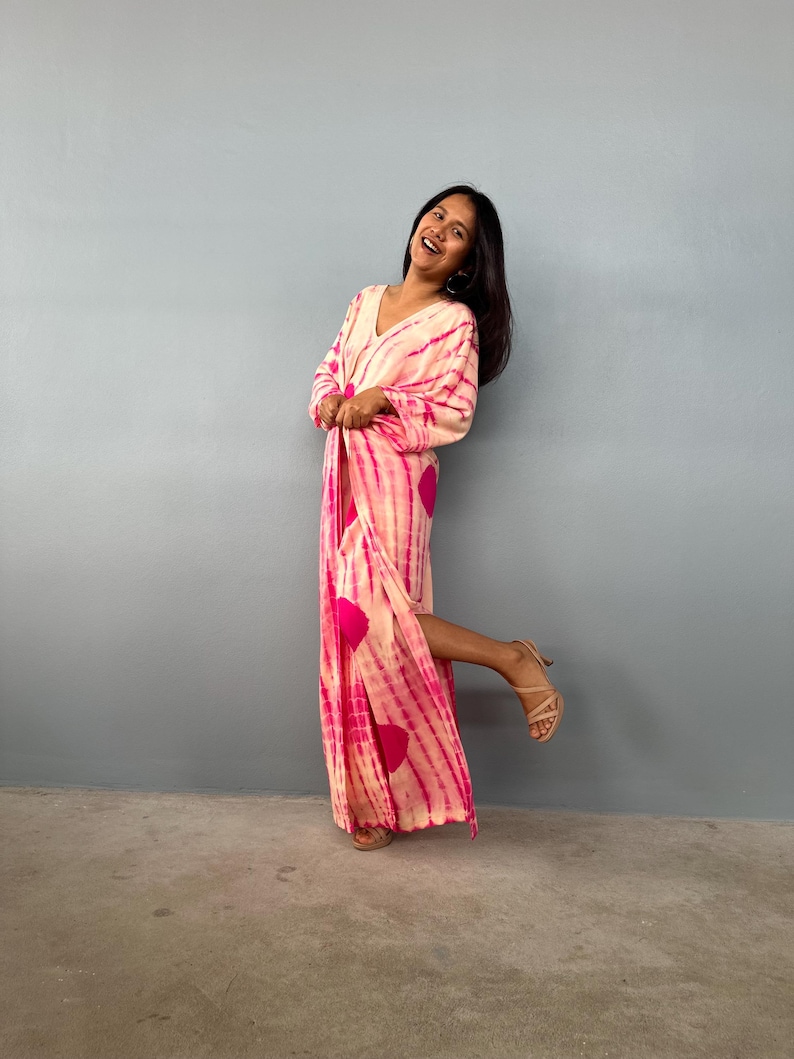 May include: A long, flowing dress with a V-neckline, featuring a tie-dye pattern in shades of pink and white. The dress has a vibrant design with large pink circles and vertical stripes. The model is wearing beige high-heeled sandals.