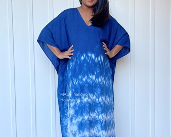 May include: A long, blue and white tie-dye kaftan with a V-neck. The kaftan is made of a soft, flowing fabric and has a relaxed fit. The fabric has a white and blue tie-dye pattern. The kaftan is worn by a person standing in front of a white wall. The text "MALA handworks Hollywood MD" is visible on the kaftan.