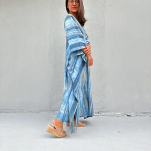 Puede incluir: Una mujer lleva una larga kaft&aacute;n a rayas azules y blancas con cuello en V y mangas 3/4. Est&aacute; de pie sobre una superficie de hormig&oacute;n y lleva sandalias marrones.