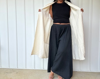 May include: A woman wearing a white, lightweight, long-sleeved top over a black sleeveless top and black wide-leg pants. She is standing in front of a white wall.