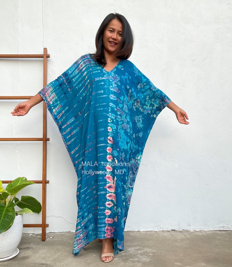 May include: A long, flowing blue and pink tie-dye kaftan with a V-neck. The kaftan has a relaxed fit and is made of a soft, comfortable fabric. The text "MALA handworks Hollywood MD" is printed on the bottom of the kaftan.