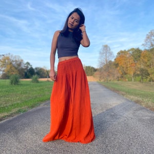 May include: A woman wearing a grey tank top and a long, flowing orange skirt. She is standing on a paved road with trees in the background.