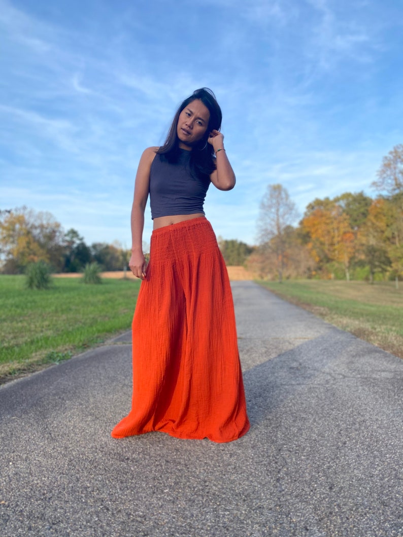 May include: A woman wearing a grey tank top and a long, flowing orange skirt. She is standing on a paved road with trees in the background.