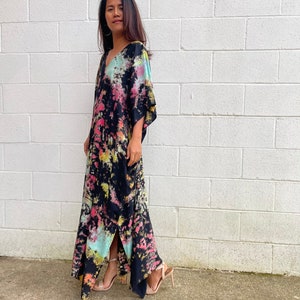May include: A long black maxi dress with a tie-dye pattern in shades of pink, blue, and green. The dress has a V-neckline and a side slit. The model is wearing the dress with nude heels.