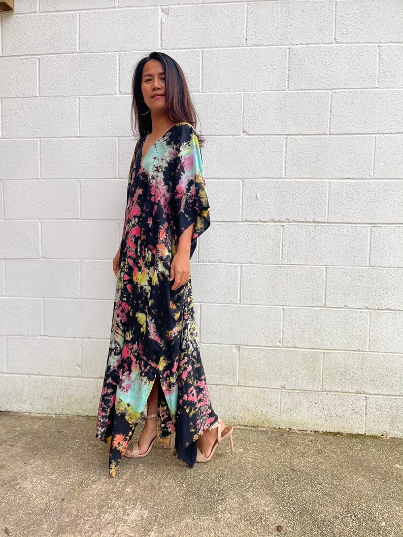 May include: A long black maxi dress with a tie-dye pattern in shades of pink, blue, and green. The dress has a V-neckline and a side slit. The model is wearing the dress with nude heels.