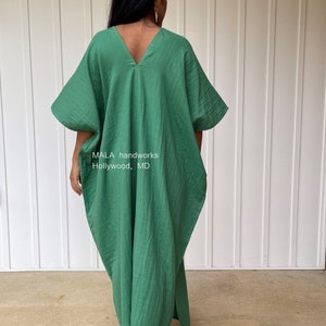 May include: A green, loose-fitting caftan dress with a V-neck and wide sleeves. The dress is made of a textured fabric and has a split on the side. The text "MALA handworks Hollywood, MD" is visible on the garment.