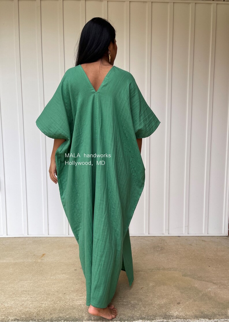 May include: A green, loose-fitting caftan dress with a V-neck and wide sleeves. The dress is made of a textured fabric and has a split on the side. The text "MALA handworks Hollywood, MD" is visible on the garment.