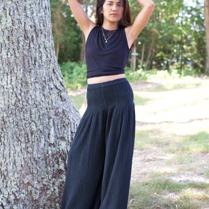 May include: A woman wearing a black sleeveless top and black wide-leg pants stands in a forest setting. She is wearing a gold necklace.