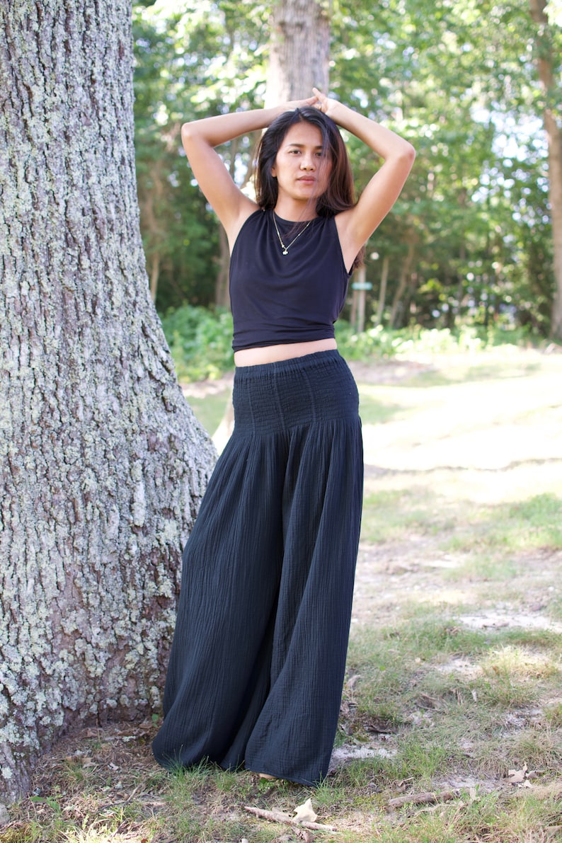 May include: A woman wearing a black sleeveless top and black wide-leg pants stands in a forest setting. She is wearing a gold necklace.