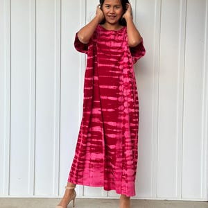 May include: A long, loose-fitting dress with a tie-dye pattern in shades of pink and red. The dress has short sleeves and a round neckline.