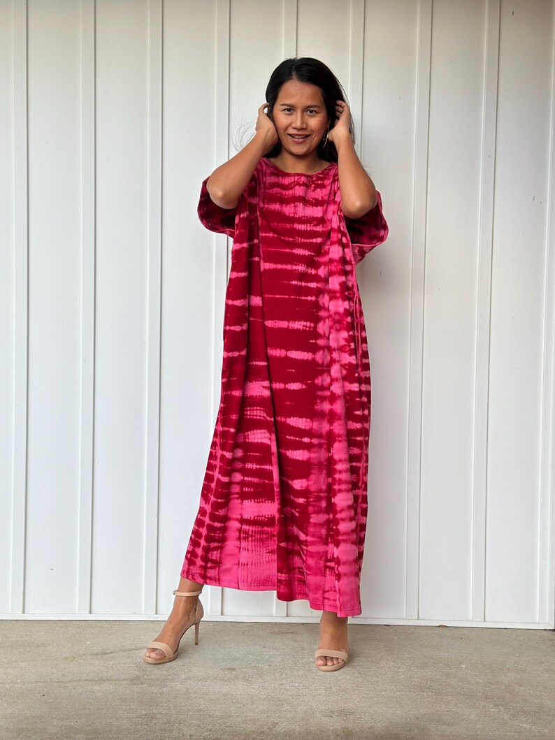 May include: A long, loose-fitting dress with a tie-dye pattern in shades of pink and red. The dress has short sleeves and a round neckline.