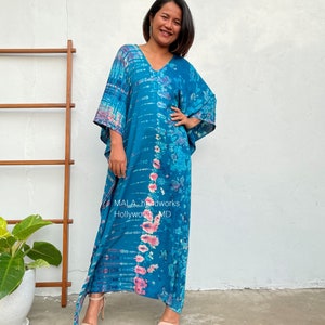 May include: A long, flowing blue and pink tie-dye kaftan with a V-neckline. The kaftan has a relaxed fit and is made of a soft, comfortable fabric. The text "MALA handworks Hollywood MD" is visible on the fabric.