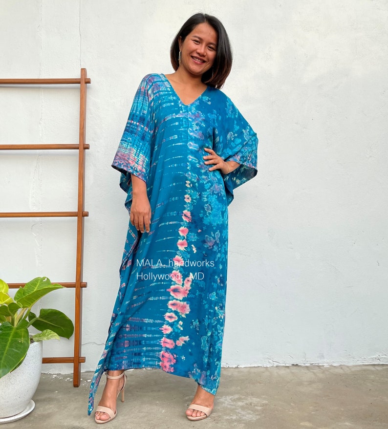 May include: A long, flowing blue and pink tie-dye kaftan with a V-neckline. The kaftan has a relaxed fit and is made of a soft, comfortable fabric. The text "MALA handworks Hollywood MD" is visible on the fabric.