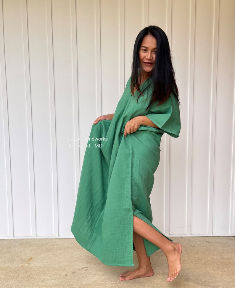 May include: A long, flowing green dress with a V-neck and wide sleeves. The dress is made of a lightweight fabric and has a high slit on one side. The text "MALA handworks Hollywood, MD" is visible on the garment.