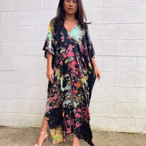 May include: A long black maxi dress with a tie-dye pattern in shades of pink, green, and yellow. The dress has a V-neck and short sleeves. The dress has a side slit and is made of a soft, flowing fabric.