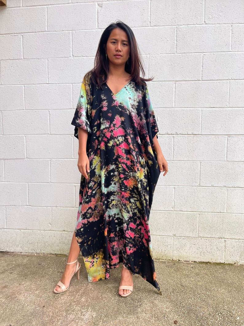 May include: A long black maxi dress with a tie-dye pattern in shades of pink, green, and yellow. The dress has a V-neck and short sleeves. The dress has a side slit and is made of a soft, flowing fabric.