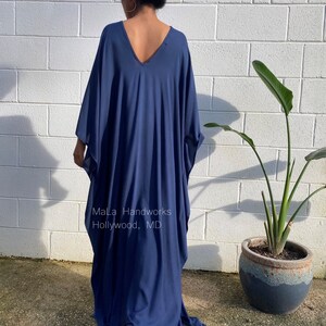 May include: A long, flowing, navy blue kaftan dress with a deep V-neckline. The dress is made of a soft, comfortable fabric and has a relaxed fit. The dress is shown on a model standing in front of a white brick wall. The text "Mala Handworks Hollywood, MD" is visible on the dress.