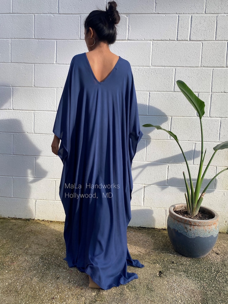 May include: A long, flowing, navy blue kaftan dress with a deep V-neckline. The dress is made of a soft, comfortable fabric and has a relaxed fit. The dress is shown on a model standing in front of a white brick wall. The text "Mala Handworks Hollywood, MD" is visible on the dress.