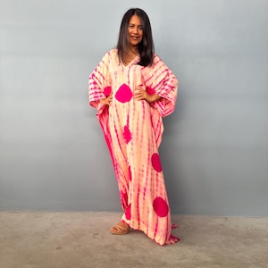 May include: A long, flowing, pink and peach kaftan dress with a tie-dye pattern and large magenta polka dots. The dress has a V-neck and wide sleeves. The person in the image is wearing brown sandals.