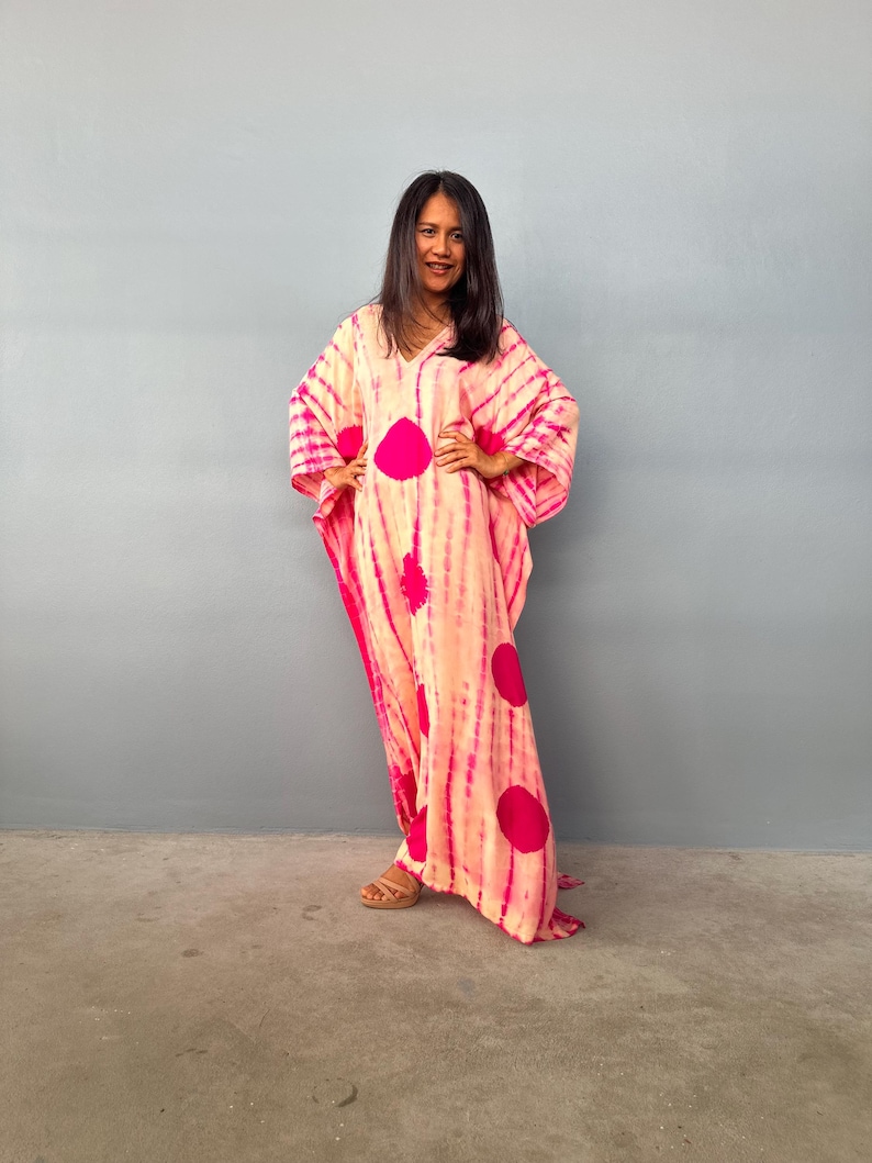 May include: A long, flowing, pink and peach kaftan dress with a tie-dye pattern and large magenta polka dots. The dress has a V-neck and wide sleeves. The person in the image is wearing brown sandals.