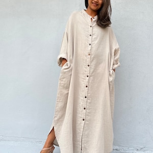 May include: A long, beige, button-down dress with a relaxed fit and side slits. The dress has a collar and long sleeves.