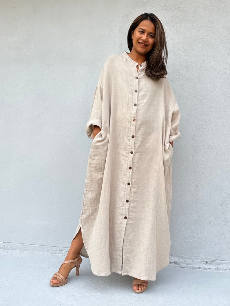 May include: A long, beige, button-down dress with a relaxed fit and side slits. The dress has a collar and long sleeves.