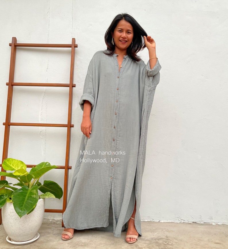 Pu&ograve; includere: Un lungo abito camicia grigio con bottoni, dalla vestibilit&agrave; rilassata. L'abito ha un colletto e maniche lunghe. La modella indossa l'abito con sandali. Il testo "MALA handworks Hollywood, MD" &egrave; visibile sull'abito.