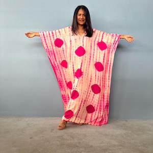 May include: A pink and cream-coloured caftan with large, vibrant pink polka dots. The caftan has a V-neck and wide sleeves. The fabric has a tie-dye effect with vertical stripes. The model is wearing brown sandals.