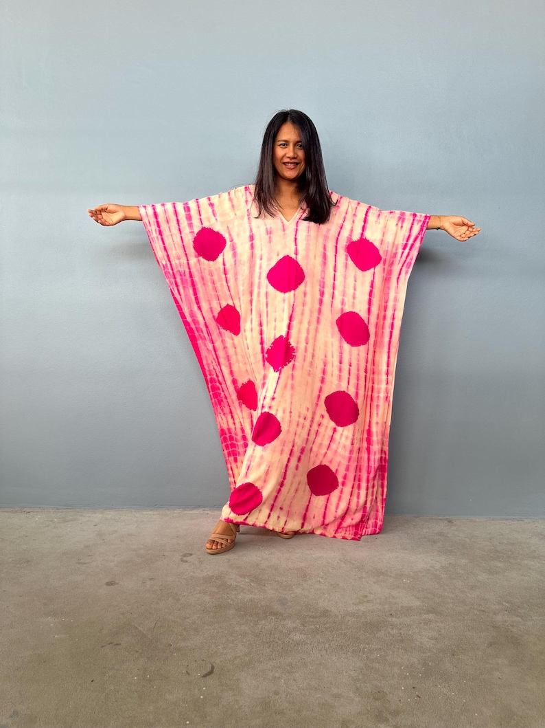 May include: A pink and cream-coloured caftan with large, vibrant pink polka dots. The caftan has a V-neck and wide sleeves. The fabric has a tie-dye effect with vertical stripes. The model is wearing brown sandals.