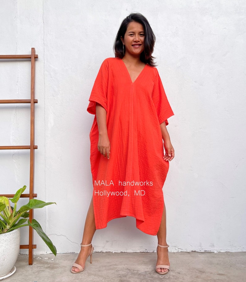 May include: A woman wearing a bright orange, loose-fitting, long-sleeved dress with a V-neck. The dress has side slits and a textured fabric. The woman is standing in front of a white wall with a wooden ladder and a potted plant. The text "MALA handworks Hollywood, MD" is visible at the bottom of the image.