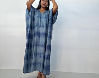 May include: A long, flowing blue tie-dye dress with a V-neck. The dress has wide sleeves and a loose fit. The fabric features a pattern of dark and light blue vertical stripes. The model is wearing tan heels.
