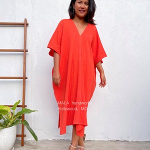 May include: A woman wearing a bright orange, loose-fitting, V-neck dress with a flowing silhouette. The dress has a slightly gathered look and is made of a soft, textured fabric. The dress is knee-length and has a relaxed fit. The woman is standing in front of a white wall with a wooden ladder and a potted plant in the background. The text "MALA handworks Hollywood, MD" is visible on the dress.