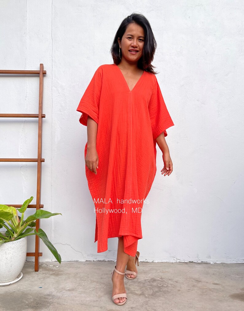 May include: A woman wearing a bright orange, loose-fitting, V-neck dress with a flowing silhouette. The dress has a slightly gathered look and is made of a soft, textured fabric. The dress is knee-length and has a relaxed fit. The woman is standing in front of a white wall with a wooden ladder and a potted plant in the background. The text "MALA handworks Hollywood, MD" is visible on the dress.
