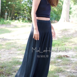 May include: A woman wearing a black sleeveless top and a long, black, flowing skirt. The skirt has a gathered waistband and is made of a lightweight fabric. The woman is standing in a grassy area with trees in the background. The text "MaLaHandworks HOLLYWOOD, MD" is visible on the skirt.
