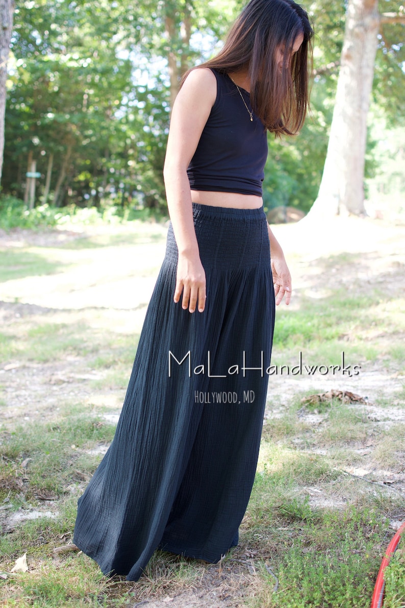 May include: A woman wearing a black sleeveless top and a long, black, flowing skirt. The skirt has a gathered waistband and is made of a lightweight fabric. The woman is standing in a grassy area with trees in the background. The text "MaLaHandworks HOLLYWOOD, MD" is visible on the skirt.
