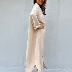 May include: A long, beige linen shirt dress with a button-up front and long sleeves. The dress has a side slit and is worn with nude heels.