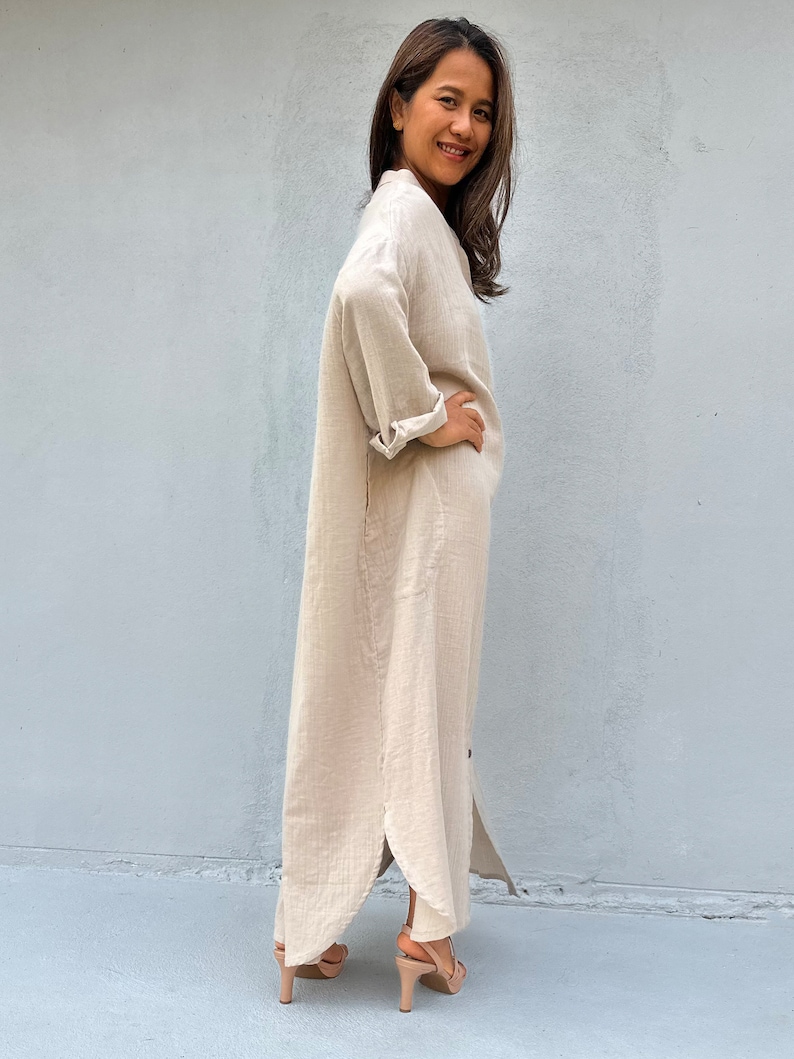 May include: A long, beige linen shirt dress with a button-up front and long sleeves. The dress has a side slit and is worn with nude heels.